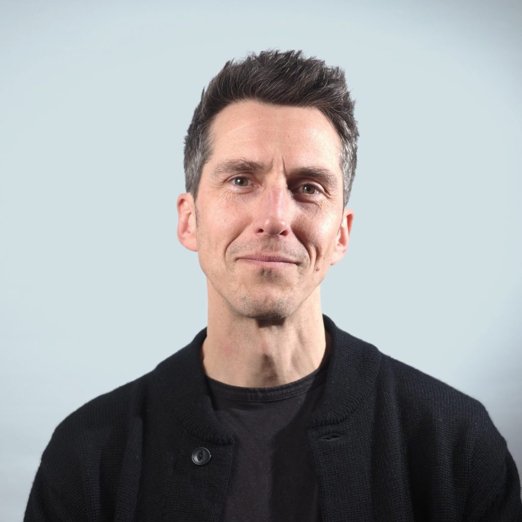 Portrait of Mathew Wilson, a middle aged white guy in black clothes, with short dark hair, and a smile that gives away how uncomfortable he feels in front of a camera.